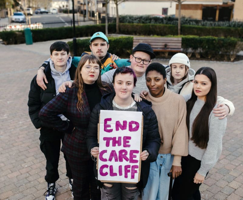 Landscape group of young people holding end the care cliff sign, looking into camera with neutral faces
