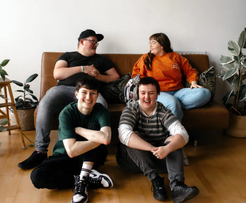 Group of young people laughing together, two sat on floor, two sat on sofa
