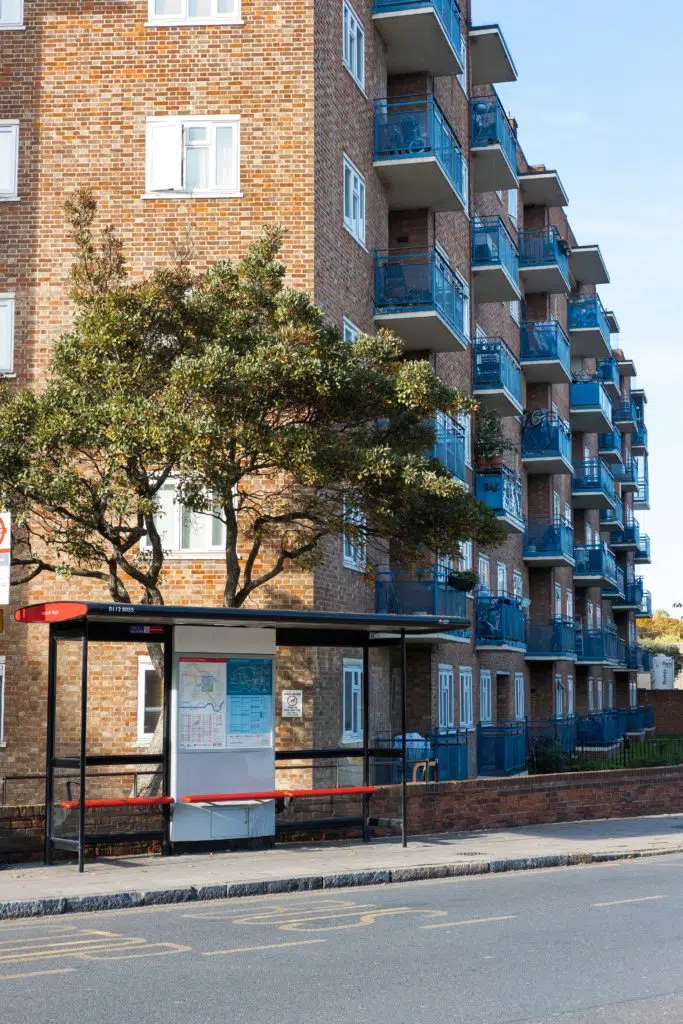 A block of flats in the background against a blue sky. In the foreground, there's a tree, a London bus stop and a road.