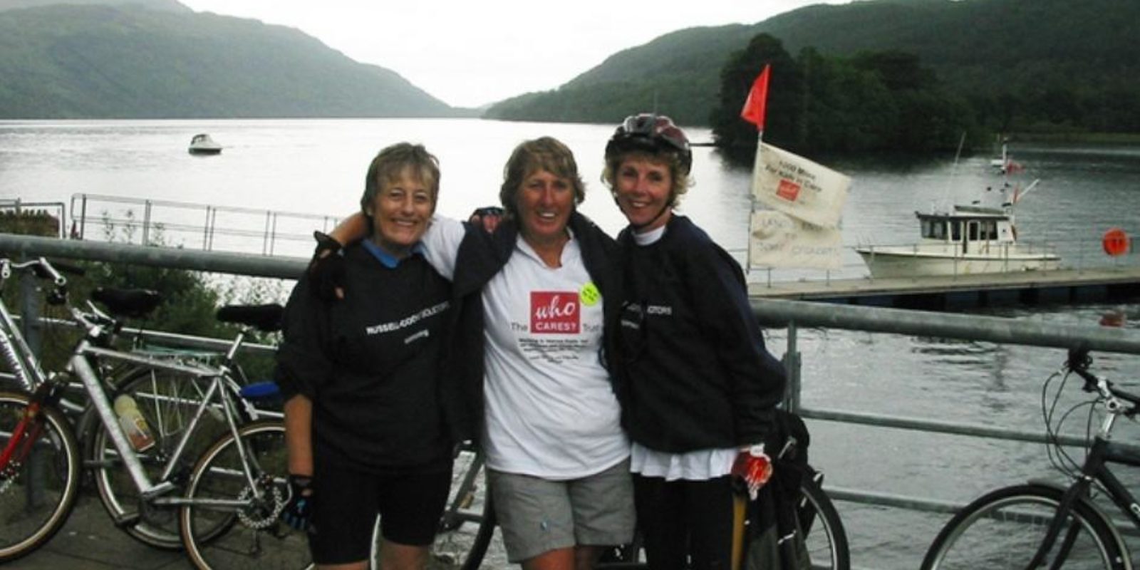 Pip, Gill and a friend stand in front of a lake with their bikes