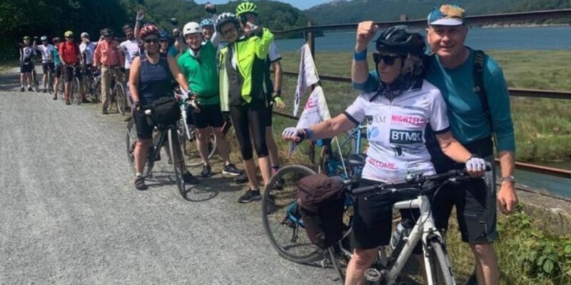 Gill and David standing in front of a line of riders on the edge of a lake
