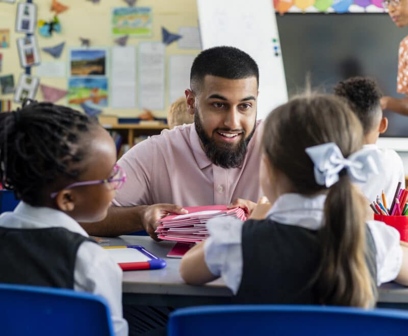Primary school students sitting in a classroom being taught by a teacher who is crouched in front of them.
