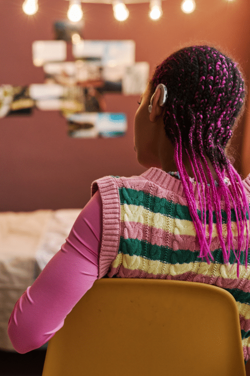 Back view of black teenage girl wearing hearing aid with pink hair in braids in cozy home interior, copy space