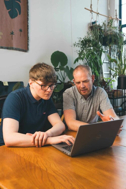 Two people looking at a laptop, sat at a desk in an office.