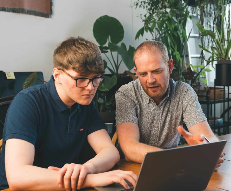Two people looking at a laptop, sat at a desk in an office.