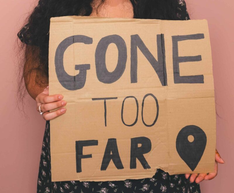 Young women with long dark black hair in a black dress with white pattern holding a cardboard placard with the word 'Gone Too Far' and a map pin.