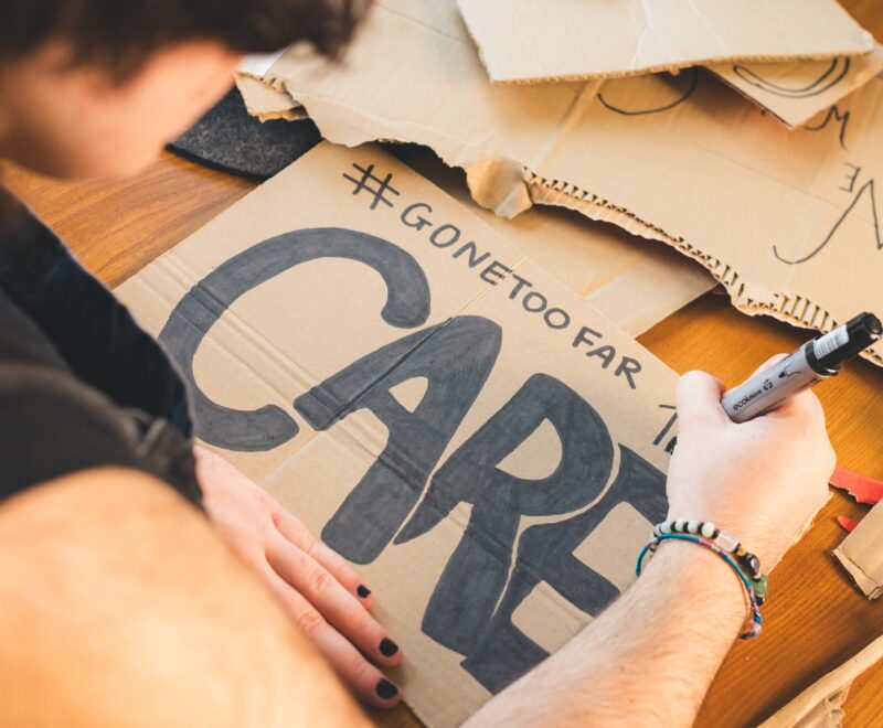 An arm holding a pen, writing the word CARE on brown cardboard.
