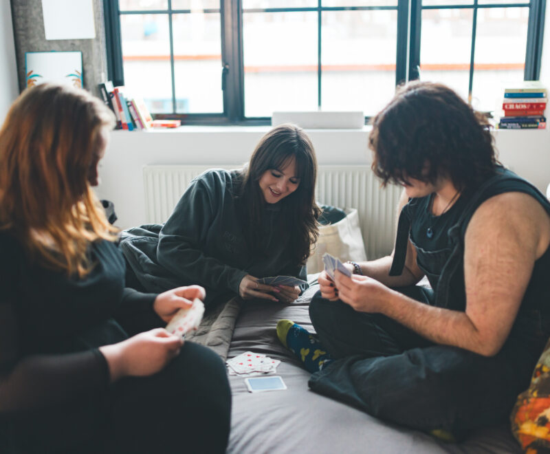 Three young people sat on a bed playing cards.