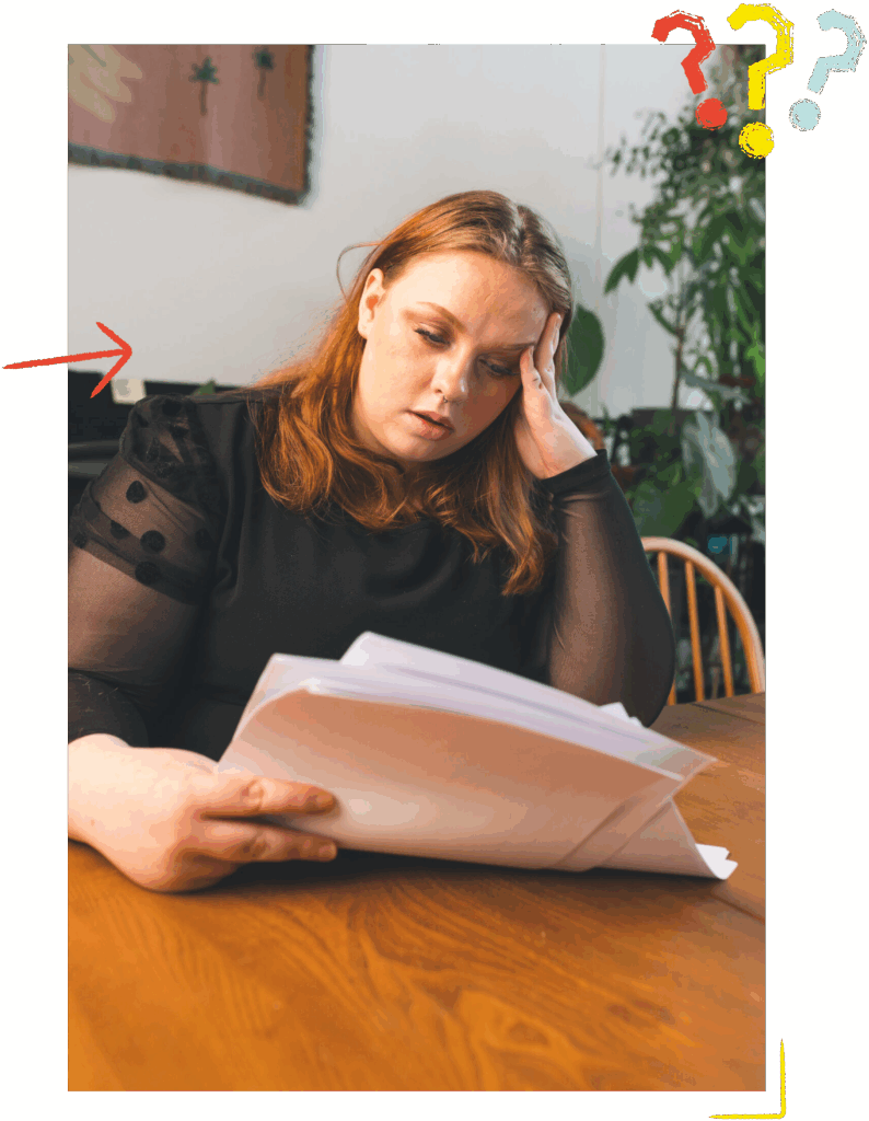 Young person with auburn hair and black top sat at a table looking stressed while reading some papers.
