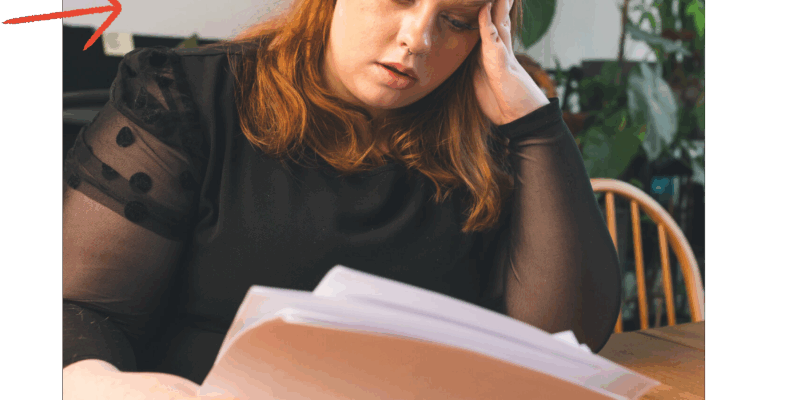 Young person with auburn hair and black top sat at a table looking stressed while reading some papers.